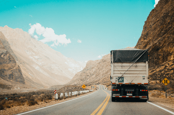 Shipping truck driving down road towards mountain range on a sunny day.